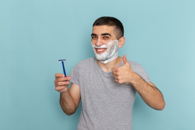 Vista frontal del varón joven en camiseta gris sosteniendo la navaja y sonriendo en la barba de color azul hielo macho de afeitar con espuma