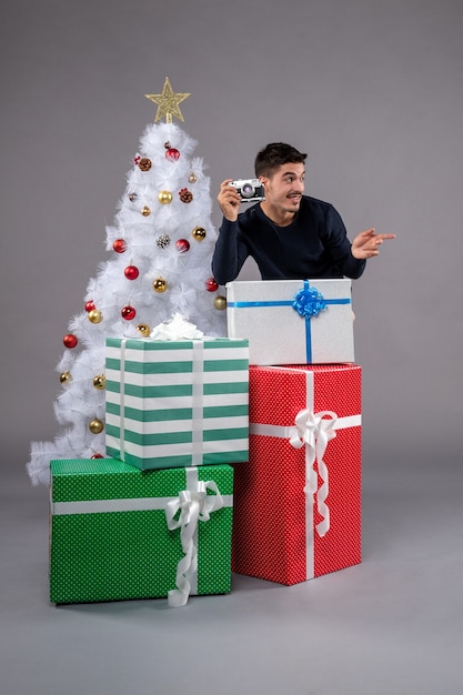 Vista frontal del varón joven con regalos y cámara en escritorio gris vacaciones año nuevo navidad