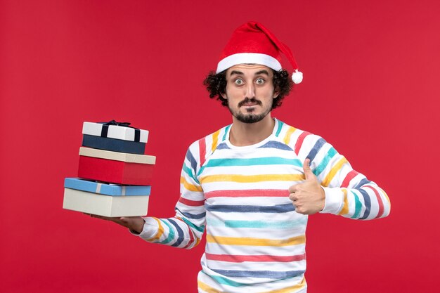 Vista frontal varón joven sosteniendo regalos navideños en la pared roja vacaciones emociones de año nuevo
