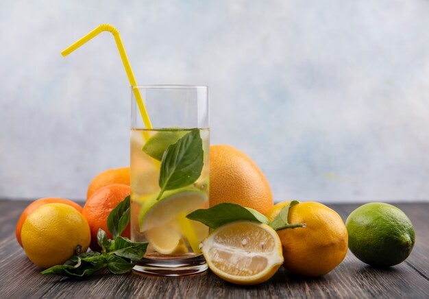 Vista frontal vaso de agua con rodajas de lima limón y menta con amarillo paja y pomelo con naranjas sobre fondo de madera