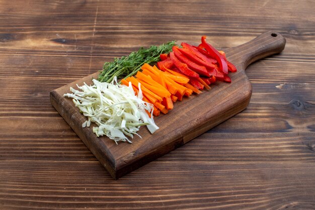 Vista frontal de verduras en rodajas, repollo, zanahoria y pimiento en la superficie marrón de la tabla de cortar