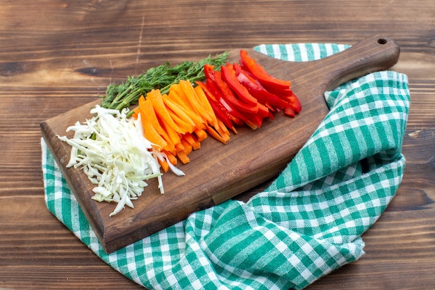 Vista frontal de verduras en rodajas, repollo, zanahoria y pimiento en la superficie marrón de la tabla de cortar