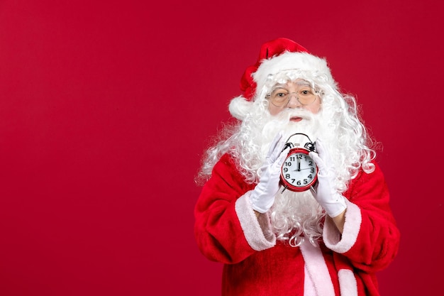 Vista frontal del viejo santa claus en traje rojo con reloj en la pared roja