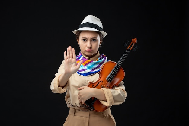 Vista frontal violinista femenina con sombrero sosteniendo violín en la pared oscura melodía instrumento música emoción concierto tocar actuación mujer