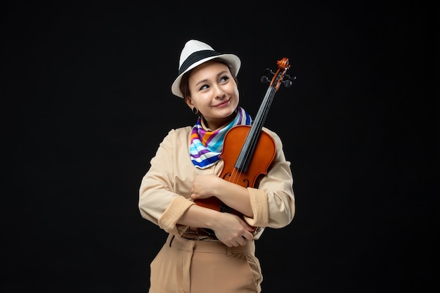 Foto gratuita vista frontal violinista femenina con sombrero sosteniendo el violín en la pared oscura melodía interpretación de instrumentos música emoción mujer concierto toca