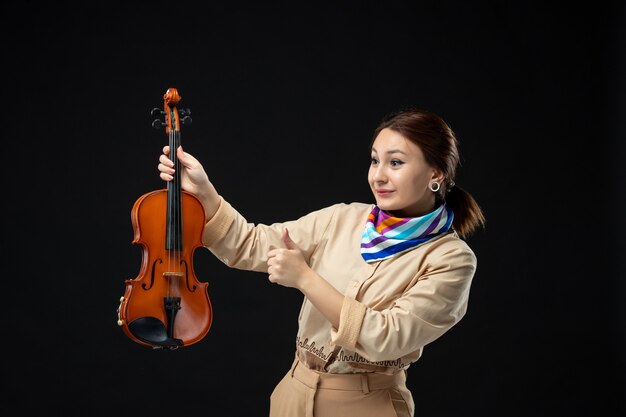 Vista frontal violinista sosteniendo su violín en la pared oscura melodía instrumento musical mujer concierto interpretación juega emoción