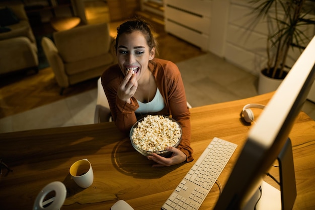 Vista de gran angular de una mujer sonriente viendo algo en Internet mientras come palomitas de maíz por la noche en casa