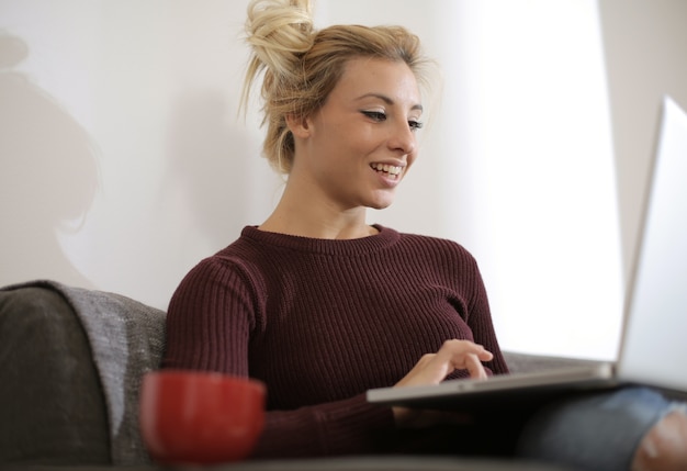 Vista de una hermosa mujer caucásica sentada en el sofá mientras trabaja felizmente en la computadora portátil