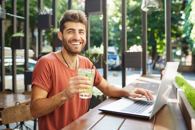 Vista del hombre barbudo caucásico alegre usando computadora portátil sonriendo con dientes, navegando por internet sentado en la mesa de madera en la cafetería de verano y bebiendo limonada.