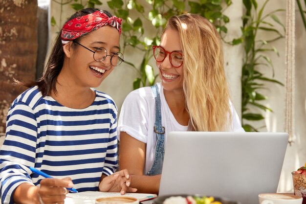 Vista horizontal de alegres mujeres de raza mixta que escriben un artículo sobre un trabajo distante, aprenden el idioma por Internet
