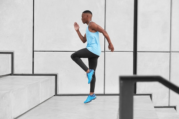 La vista horizontal del atleta masculino vestido con ropa deportiva, tiene cardio corriendo escaleras arriba se prepara para trotar a larga distancia hace escalones o saltos altos. Hombre afroamericano en camiseta, leggings, zapatillas