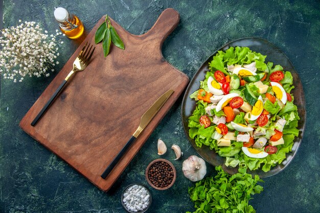 Vista horizontal de una deliciosa ensalada con muchos ingredientes frescos, especias, botella de aceite, ajos, cubiertos, sobre tabla de cortar de madera