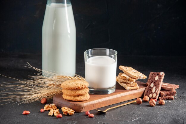 Vista horizontal de deliciosa leche en vaso y botella, cuchara dorada, galletas apiladas en bandeja de madera, flor de espigas sobre superficie oscura