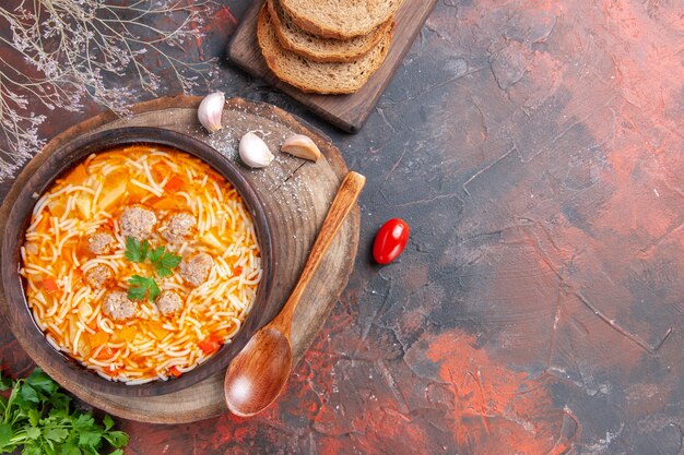 Vista horizontal de la deliciosa sopa de fideos con pollo en la tabla de cortar de madera, cuchara de verduras, ajo y tomate sobre fondo oscuro