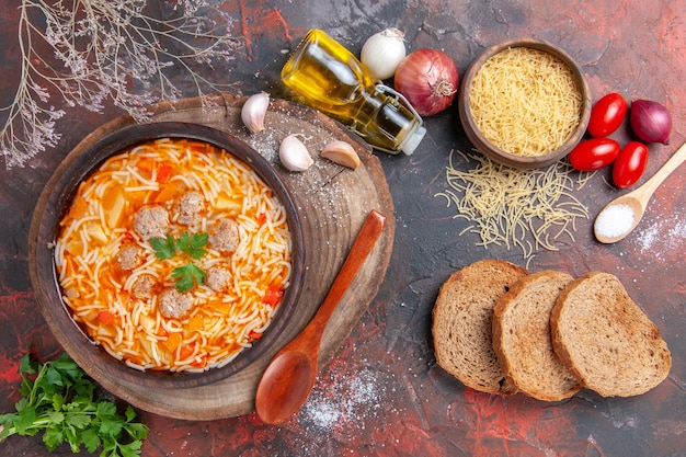 Vista horizontal de la deliciosa sopa de fideos con pollo en la tabla de cortar de madera verdes cuchara ajo tomate y rebanadas de pan sobre fondo oscuro