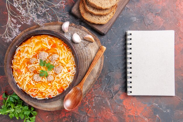 Vista horizontal de la deliciosa sopa de fideos con pollo en la tabla de cortar de madera verduras cuchara ajo tomate y cuaderno sobre fondo oscuro
