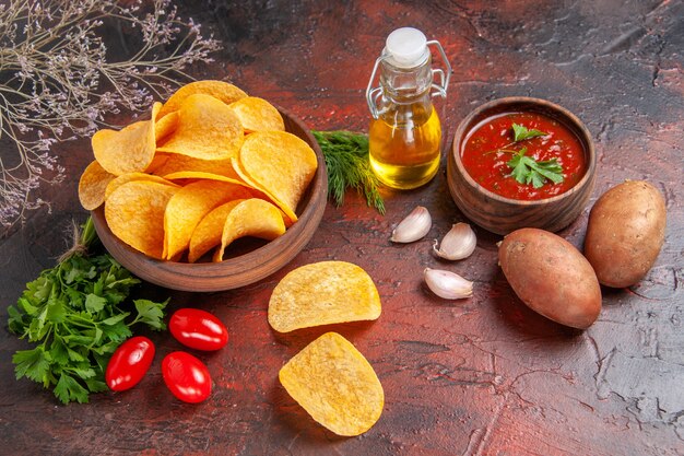 Vista horizontal de deliciosas patatas fritas crujientes caseras en un pequeño tazón marrón botella de aceite de patatas tomates verdes ajo y salsa de tomate sobre fondo oscuro