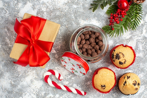 Vista horizontal de deliciosos cupcakes pequeños y chocolate en una olla de vidrio y ramas de abeto junto al regalo con cinta roja sobre la superficie del hielo