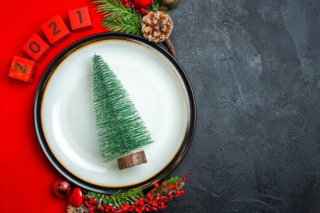 Vista horizontal del fondo de año nuevo con accesorios de decoración de plato de cena de árbol de Navidad ramas de abeto y números en una servilleta roja sobre una mesa negra