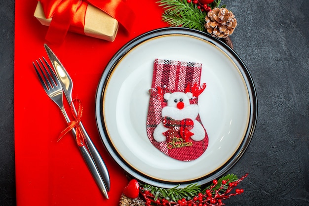 Vista horizontal de fondo de año nuevo con calcetín en plato de cena cubiertos accesorios de decoración ramas de abeto junto a un regalo en una servilleta roja