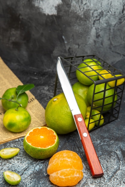 Vista horizontal de frutas cítricas frescas con hojas caídas de una canasta negra cortada por la mitad y un cuchillo en un periódico sobre fondo gris