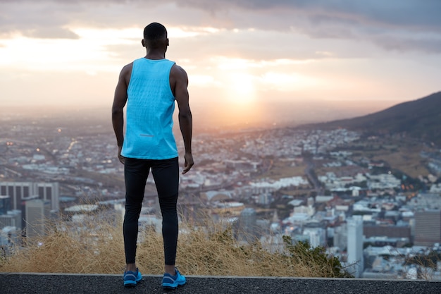 Vista horizontal de un hombre atractivo vestido con ropa casual, tiene una carrera activa cerca de las montañas, retrocede, mira atentamente el hermoso amanecer al amanecer, respira aire fresco, siente libertad, espacio libre