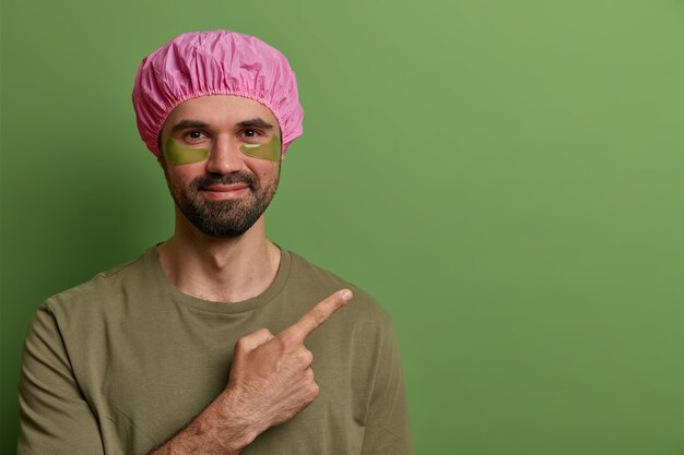 La vista horizontal del hombre satisfecho sin afeitar usa gorro de baño rosa, camiseta informal, tiene tratamiento para la piel de los ojos, aplica almohadillas de colágeno para reducir las líneas finas, señala el espacio en blanco, anuncia algún producto