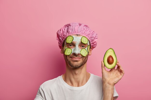 Vista horizontal del joven caucásico complacido muestra la mitad del aguacate, disfruta de una mascarilla de arcilla nutritiva en la cara, tiene rastrojo, vestido con una camiseta blanca