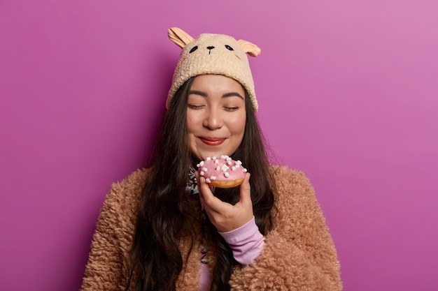 Vista horizontal de una mujer encantadora con expresión complacida, cierra los ojos y huele una deliciosa rosquilla, viste ropa de invierno, aislada sobre una pared lila