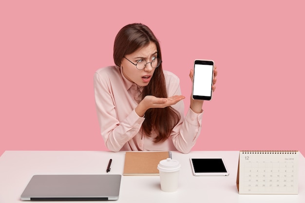 La vista horizontal de la mujer frustrada demuestra algo en la pantalla vacía del teléfono móvil