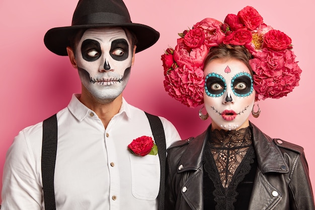 Vista horizontal de la pareja sorprendida con cara de miedo, calaveras de azúcar pintadas y sonrisas, celebran juntos el carnaval popular