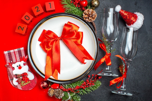 Vista horizontal de regalo con cinta roja, platos de cena, accesorios de decoración, ramas de abeto, calcetín de Navidad, copas de vidrio, sombrero de santa claus en la mesa oscura