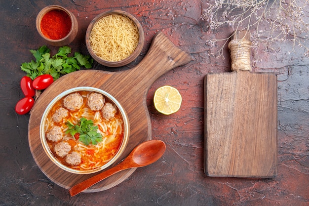 Vista horizontal de sopa de albóndigas de tomate con fideos en un recipiente marrón diferentes especias y tabla de cortar de madera sobre fondo oscuro