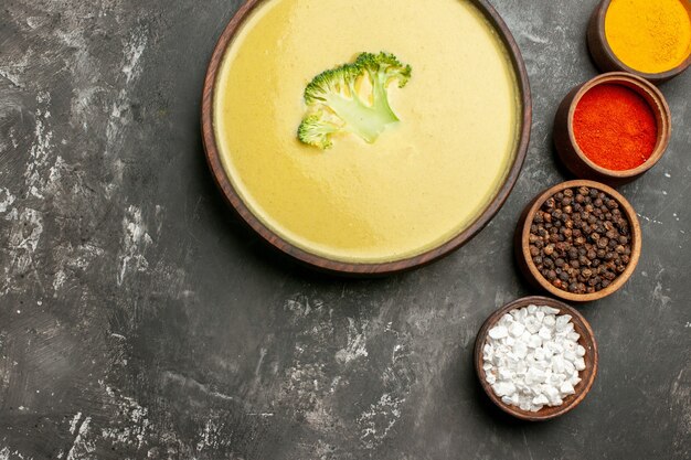 Vista horizontal de sopa cremosa de brócoli en un recipiente marrón y diferentes especias en mesa gris