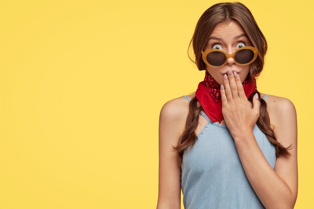 La vista de la imagen de la mujer de cabello oscuro sorprendida cubre la boca con la mano, usa tonos de moda, pañuelo rojo, mira con ojos saltones