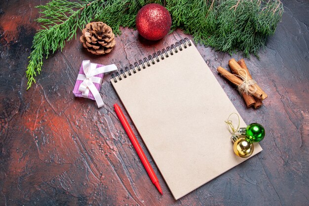 Vista inferior bolígrafo rojo un cuaderno ramas de pino árbol de navidad juguetes de bolas palitos de canela sobre fondo rojo oscuro foto de navidad