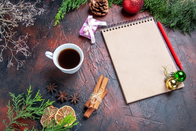 Vista inferior bolígrafo rojo un cuaderno ramas de pino árbol de navidad juguetes de bolas palitos de canela una taza de té sobre fondo rojo oscuro