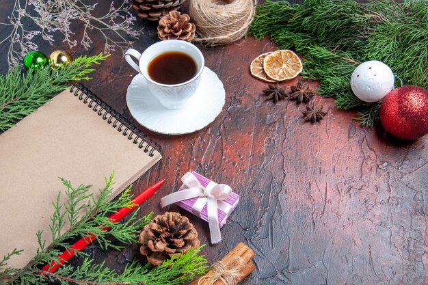 Vista inferior bolígrafo rojo un cuaderno ramas de pino árbol de navidad juguetes de bolas y regalo canela anís hilo de paja una taza de té sobre fondo rojo oscuro espacio libre