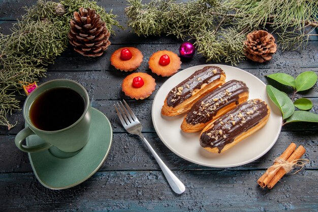 Vista inferior canutillos de chocolate en plato ovalado blanco ramas de abeto y conos juguetes de navidad un tenedor canela una taza de té y cupcakes en la mesa de madera oscura