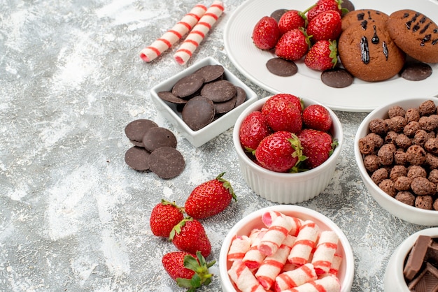 Vista inferior cercana galletas fresas y chocolates redondos en el plato ovalado blanco cuencos con dulces fresas chocolates cereales en el lado derecho de la mesa gris-blanca con espacio libre