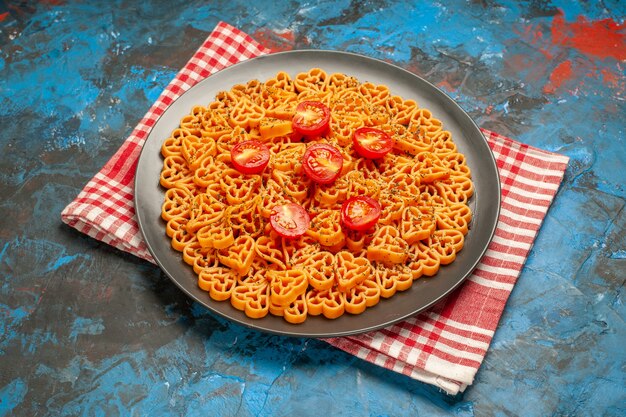 Vista inferior corazones de pasta italiana cortan tomates cherry en un plato sobre una toalla de cocina a cuadros rojo blanco sobre la mesa azul