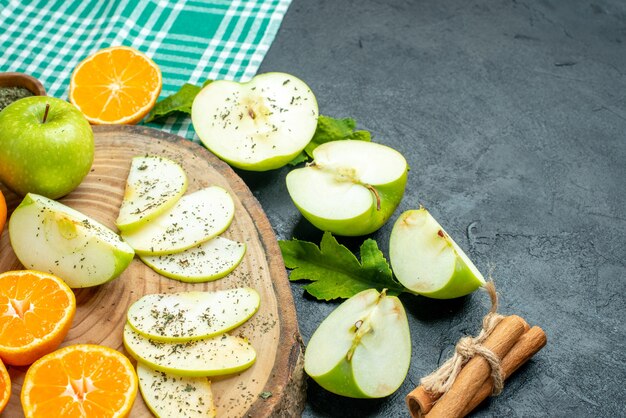 Vista inferior cortar manzanas y mandarinas sobre tablero de madera, palitos de canela atados con una cuerda sobre un mantel verde en el lugar libre de la mesa oscura