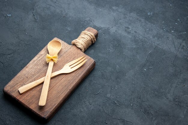 Vista inferior cruzada cuchara de madera y tenedor sobre tabla de servir de madera sobre una superficie oscura con espacio libre