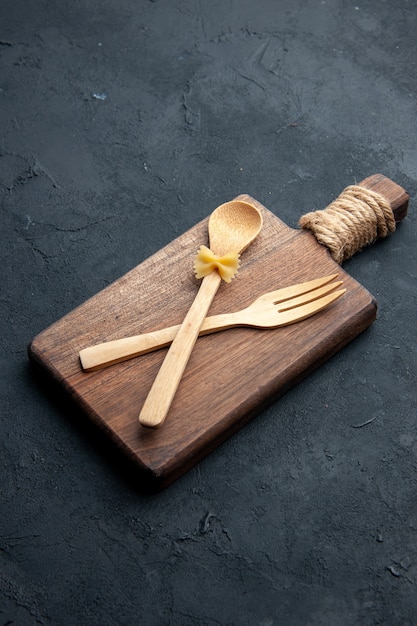 Vista inferior cruzada cuchara de madera y tenedor sobre tabla de servir de madera sobre superficie oscura