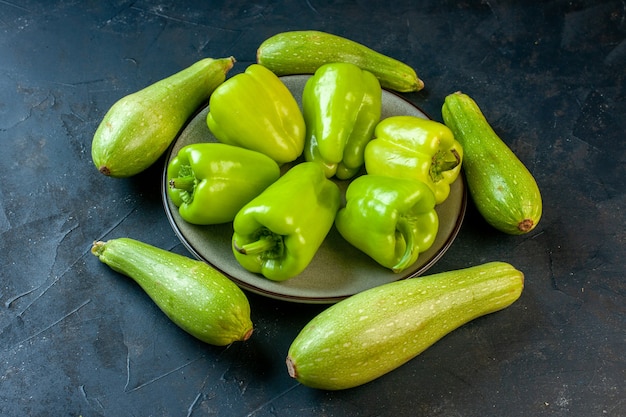 Vista inferior de diferentes tamaños de calabacín alrededor de pimientos verdes en placa redonda gris sobre superficie oscura