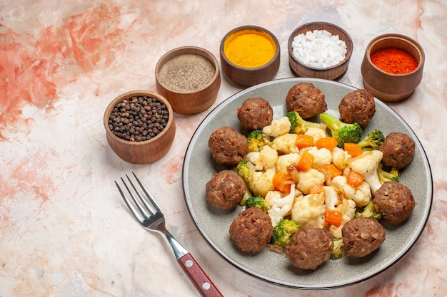 Foto gratuita vista inferior ensalada de brócoli y coliflor y albóndigas en un plato diferentes especias en tazones pequeños un tenedor sobre fondo desnudo aislado