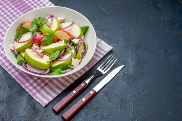 Vista inferior de la ensalada de manzana en un tazón de mantel a cuadros morado y blanco, cuchillo y tenedor en el lugar libre de la mesa oscura