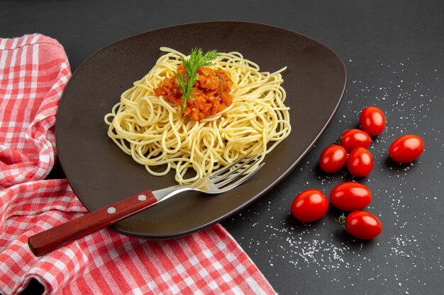 Vista inferior de espaguetis con salsa tenedor en un plato de tomates cherry, toalla de cocina a cuadros rojo blanco sobre mesa negra