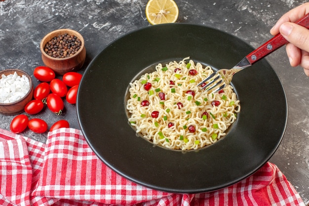 Vista inferior de fideos ramen asiáticos en plato negro horquilla en mano femenina cortada limón tomates cherry sal marina pimienta negra en tazones pequeños sobre mesa oscura