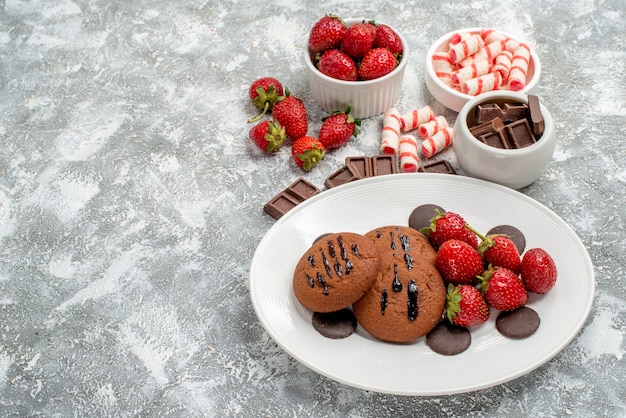 Vista inferior galletas fresas y bombones redondos en el plato blanco y cuencos de dulces fresas chocolates sobre el suelo gris-blanco